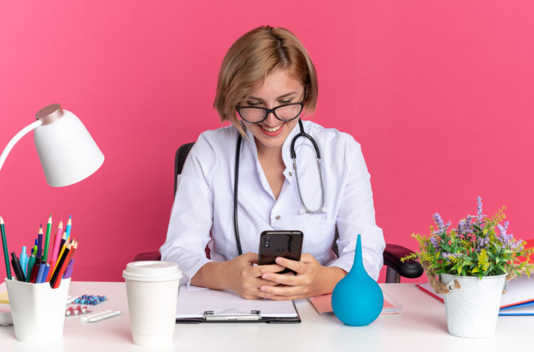 souriante jeune femme medecin portant robe medicale stethoscope lunettes est assise au bureau outils medicaux tenant regardant telephone isole fond rose
