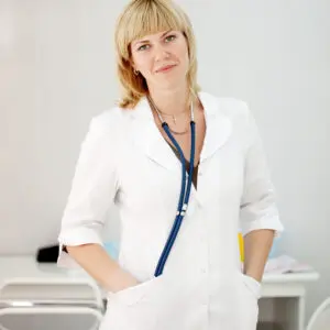 Médecin femme souriante en blouse blanche se tenant dans un cabinet médical,de Grenoble, symbolisant l'accessibilité et le professionnalisme dans les soins de santé.