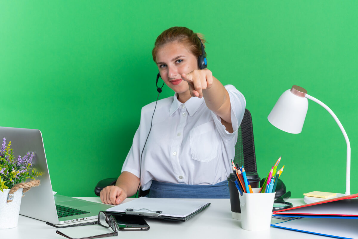 pleased young blonde call centre girl wearing headset sitting desk with work tools looking pointing camera isolated green wall