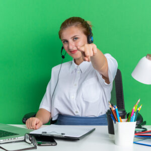 pleased young blonde call centre girl wearing headset sitting desk with work tools looking pointing camera isolated green wall