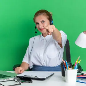 pleased young blonde call centre girl wearing headset sitting desk with work tools looking pointing camera isolated green wall